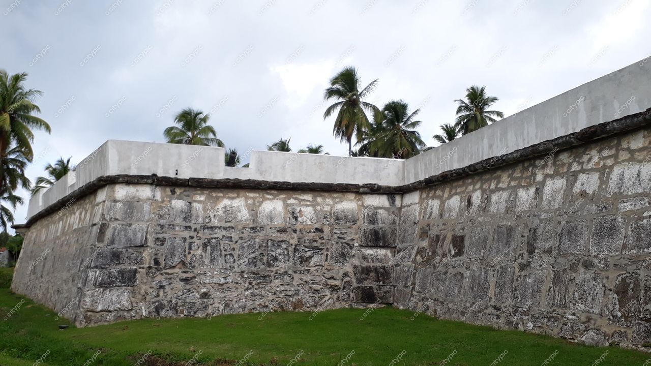 Tamandaré, Pernambuco, muralha de pedras com acabamento em cimento branco e vegetação tropical ao redor em dia nublado