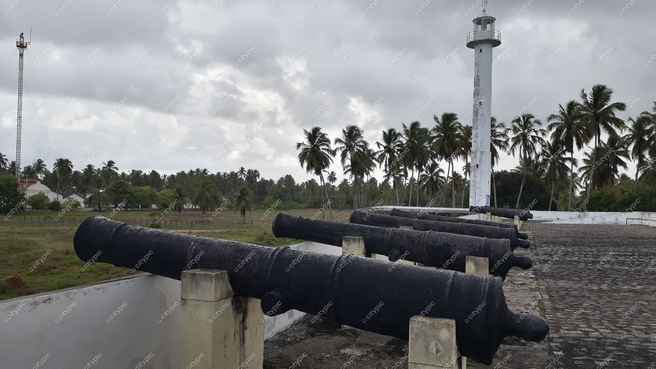 Tamandaré, Pernambuco com canhões antigos em espaço aberto e farol branco sob céu nublado e vegetação tropical