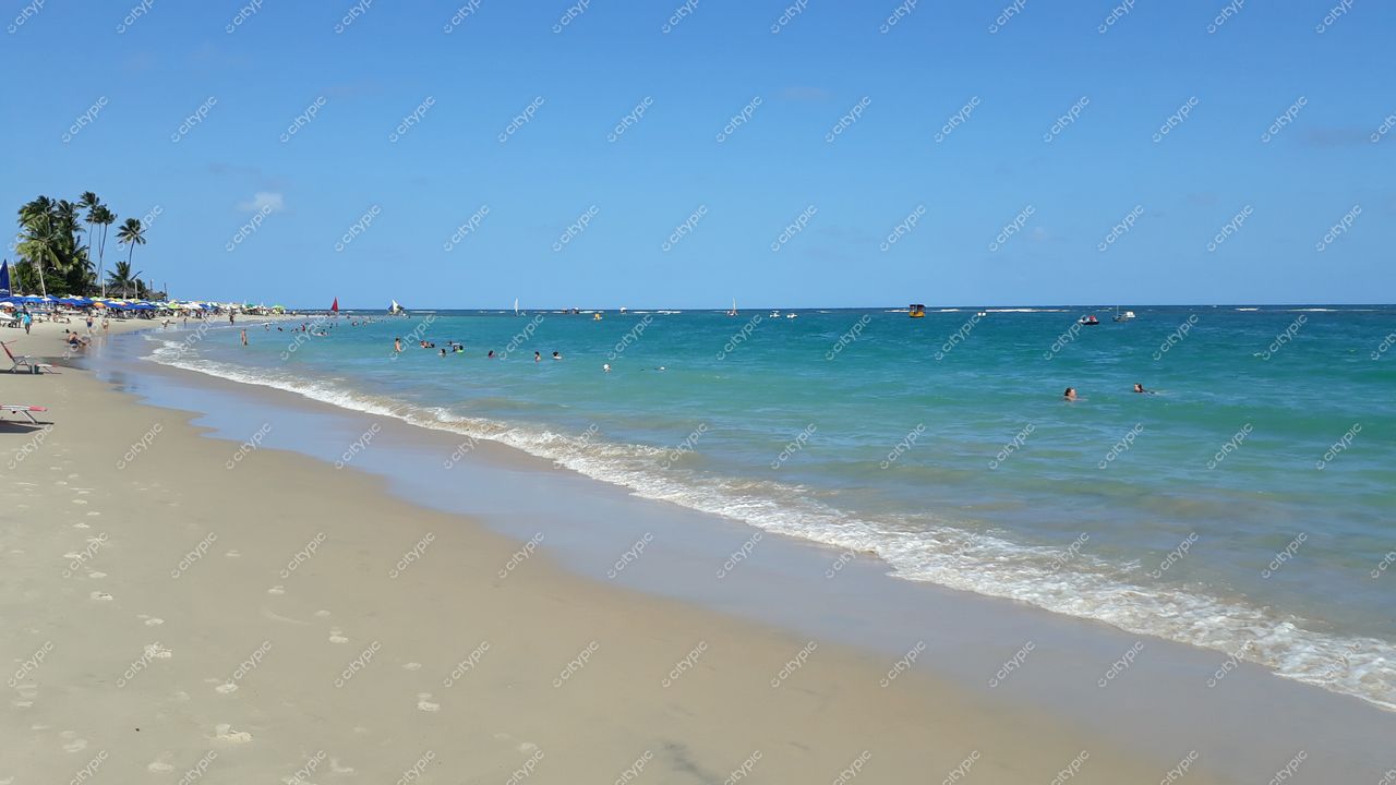 Porto de Galinhas, Pernambuco com praia cheia de pessoas sob céu azul e barcos à vela ao fundo