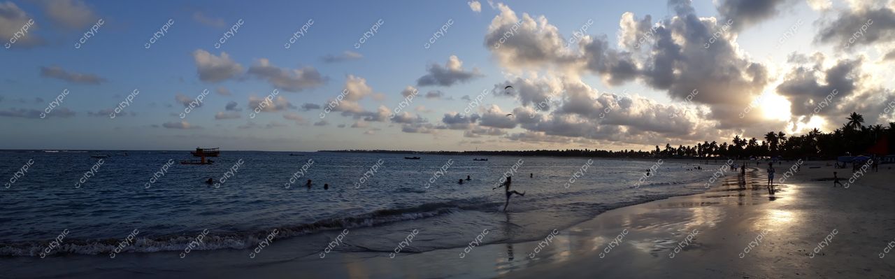 Porto de Galinhas, Pernambuco, praia ao entardecer com céu azul e laranja, pessoas na areia e palmeiras ao fundo