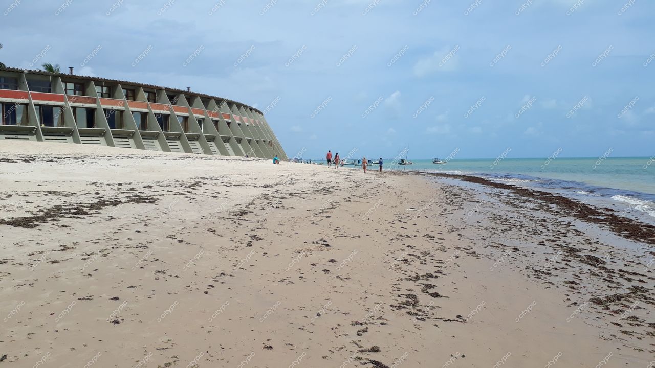 João Pessoa, Paraíba, praia com edifício distinto à direita, areia clara e pessoas caminhando sob céu parcialmente nublado