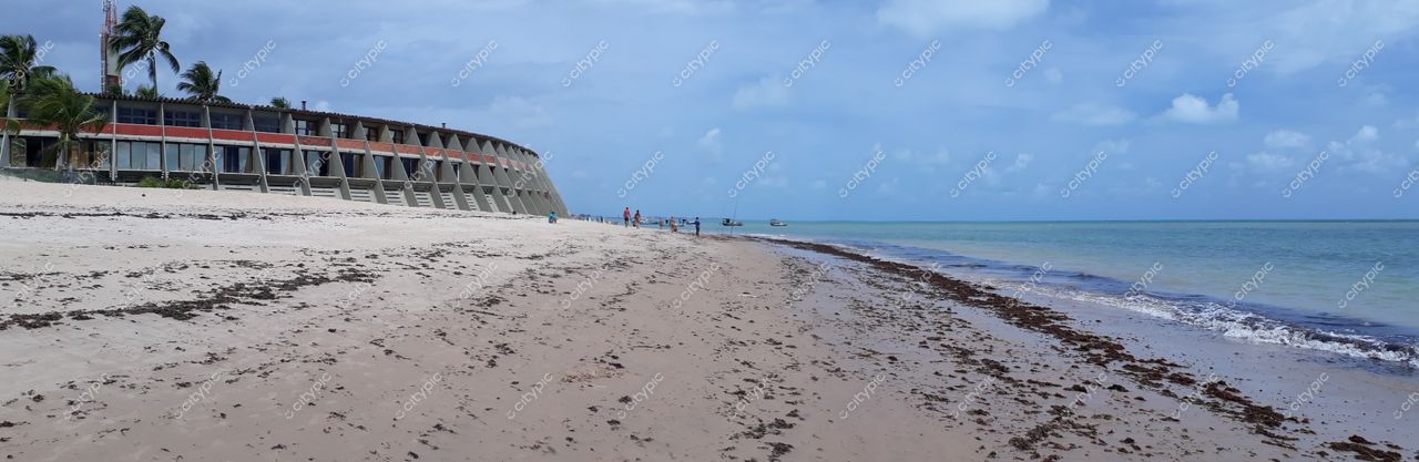 João Pessoa, Paraíba, praia com areia clara, edifício curvilíneo à beira-mar e pessoas caminhando sob céu nublado