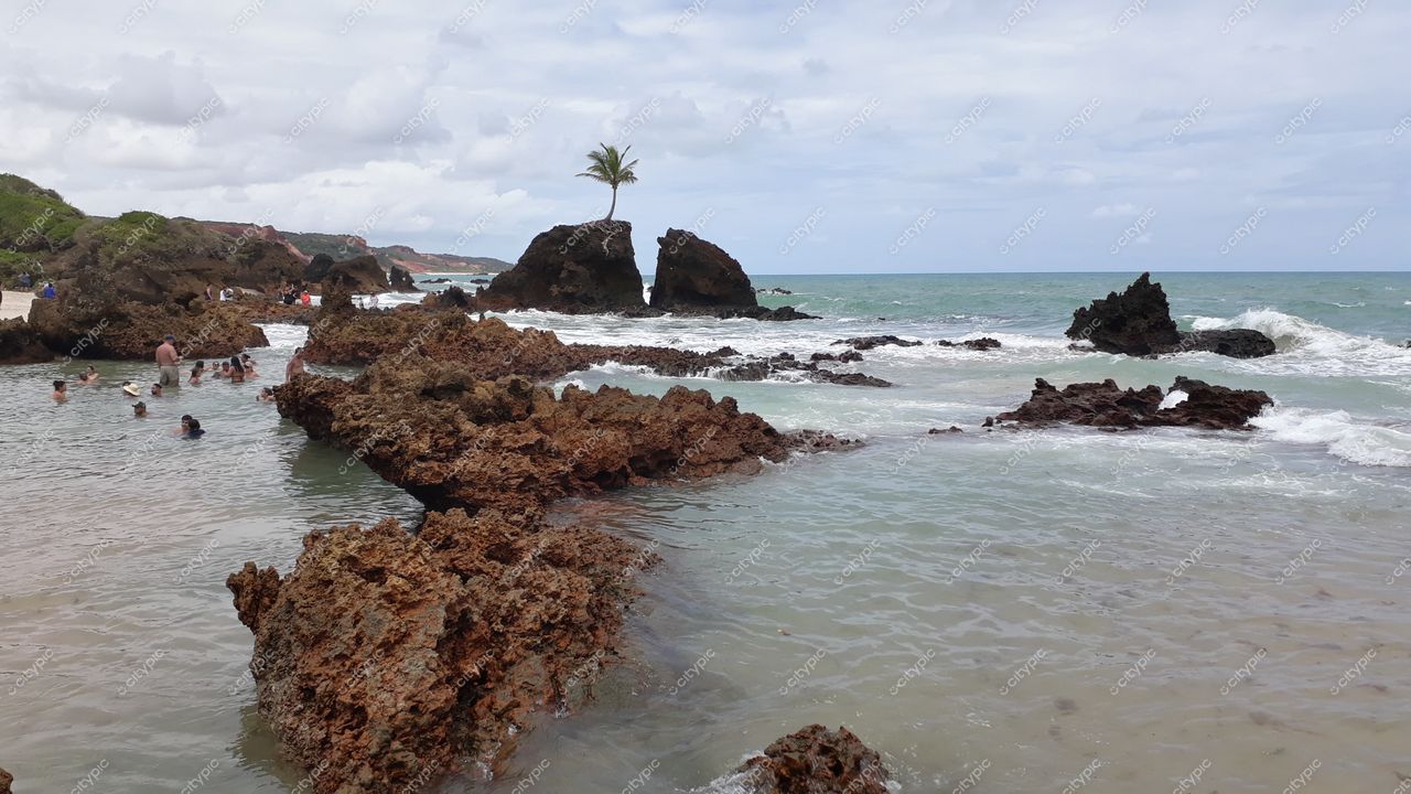 Conde, Paraíba, praia com formações rochosas expostas, vegetação ao redor e céu nublado em dia agitado