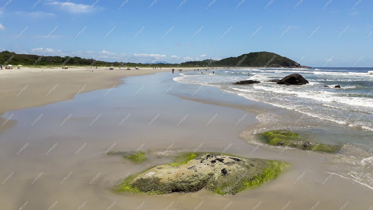 Ilha do Mel, Paraná, praia com areia clara e úmida, céu azul e formações rochosas ao fundo em dia ensolarado