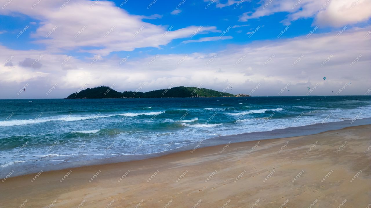 Campeche em Florianópolis com céu parcialmente nublado, ilha verdejante ao fundo e praticantes de kite surf na água