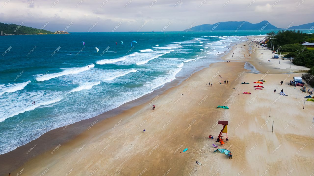 Campeche em Florianópolis com kitesurfistas nas ondas e vegetação densa sob céu nublado e montanhas ao fundo