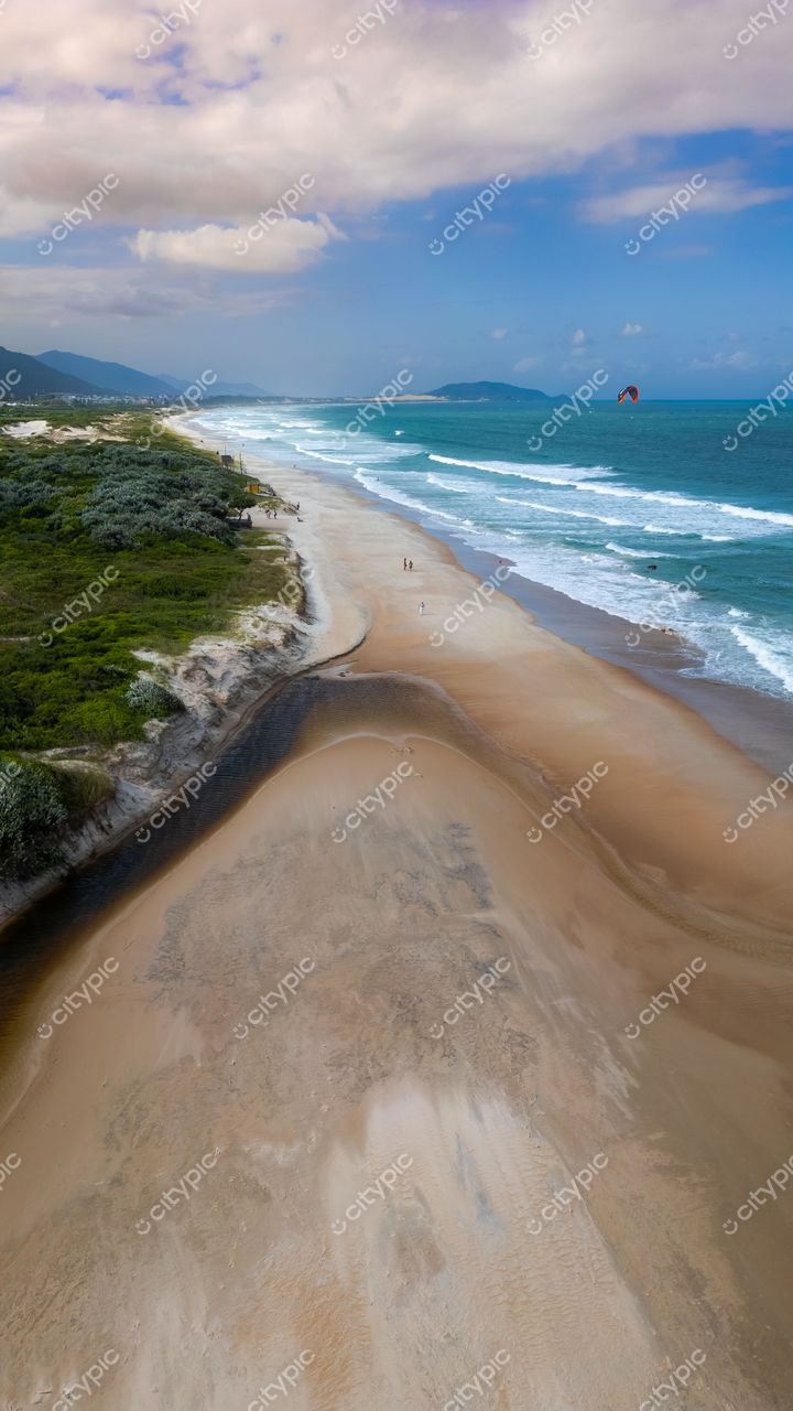 Campeche em Florianópolis com céu parcialmente nublado, vegetação rasteira e praticantes de esportes aquáticos na orla