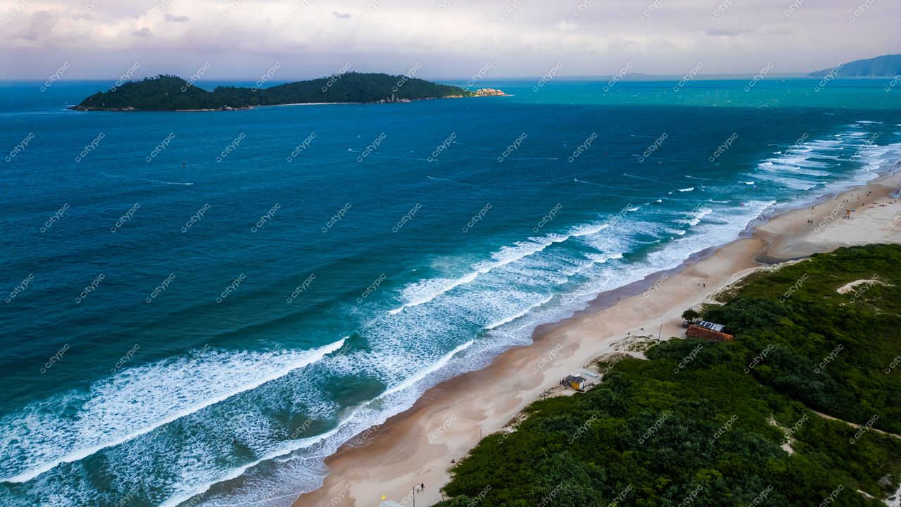 Campeche em Florianópolis com ondas suaves, vegetação densa à direita e ilhas ao fundo sob céu nublado
