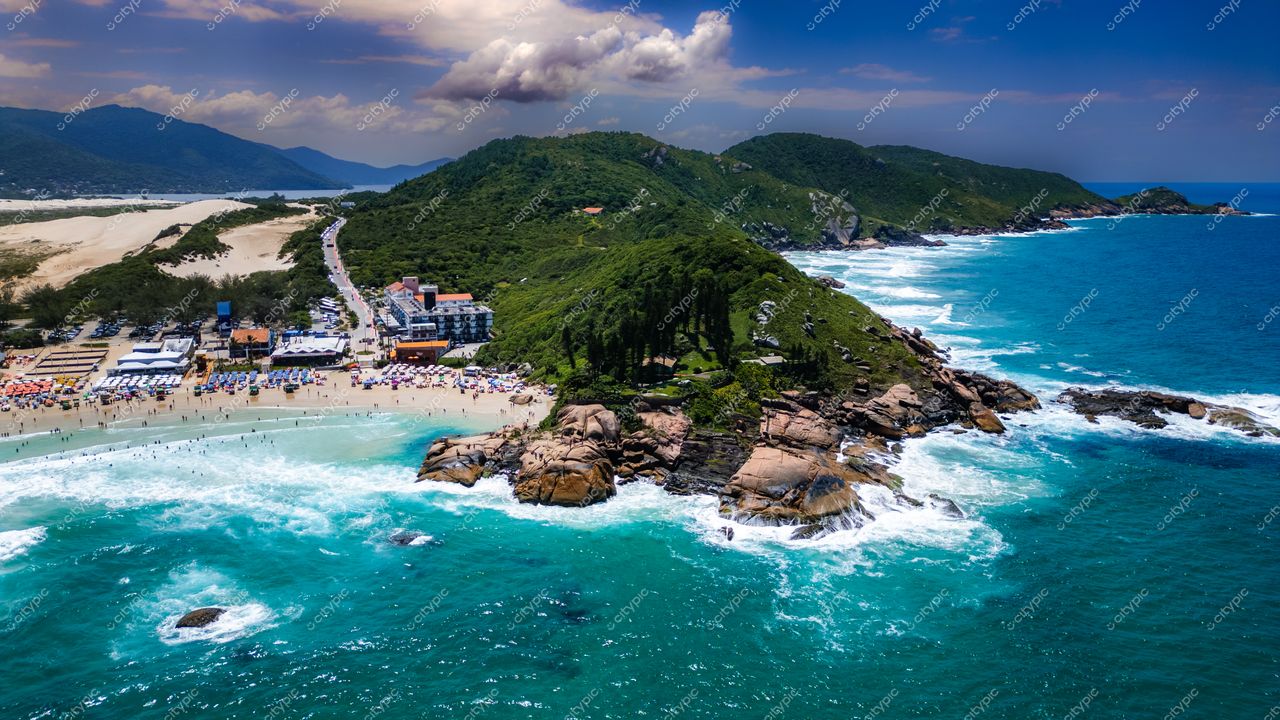Praia da Joaquina, em Florianópolis, vista aérea com águas azul-turquesa e vegetação densa ao fundo em dia claro