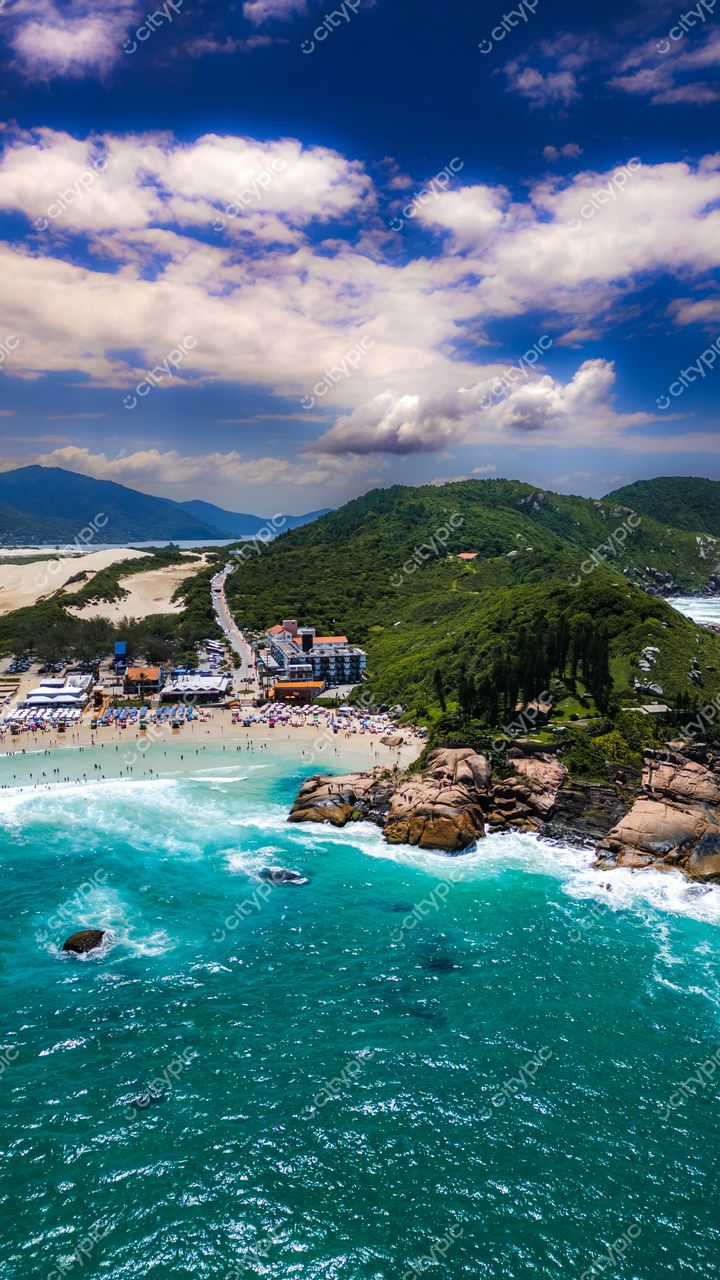 Praia da Joaquina em Florianópolis com céu azul, ondas turquesa, vegetação densa e construções à beira-mar