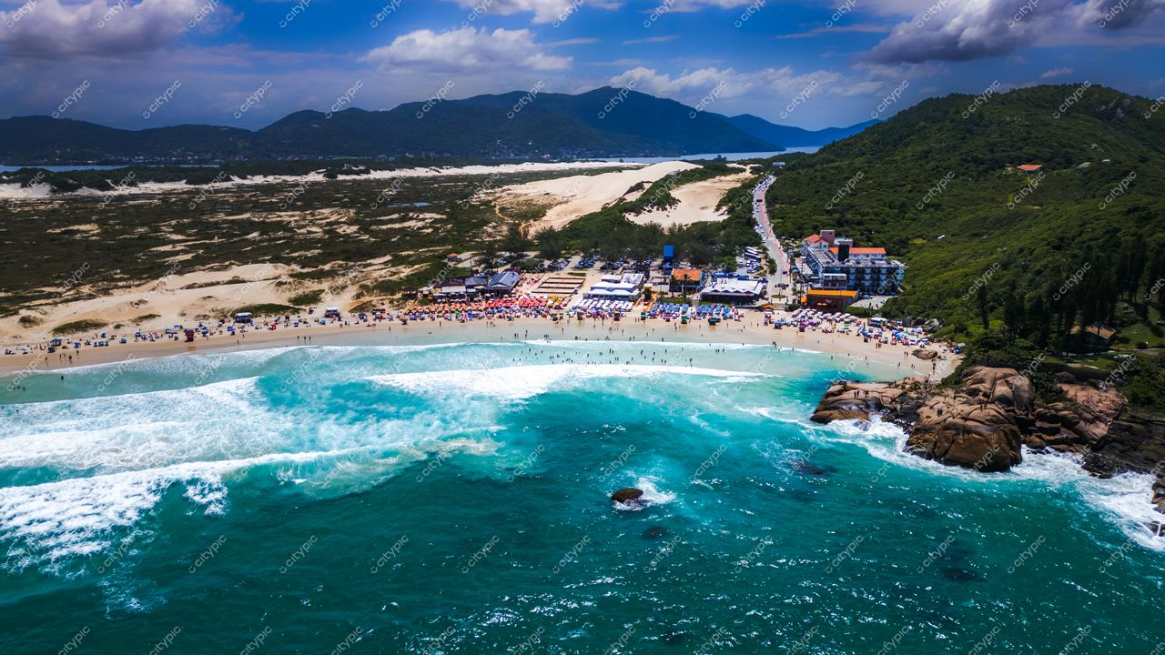 Praia da Joaquina, em Florianópolis, vista aérea com vegetação verde, hotel próximo e pessoas na areia sob céu nublado