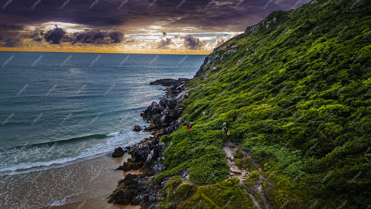 Lagoinha do Leste em Florianópolis com encosta verdejante, céu nublado e pessoas na trilha ao crepúsculo