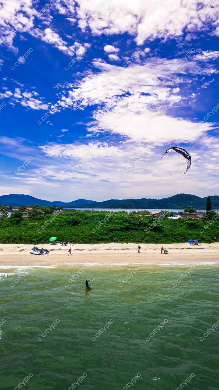 Praia da Daniela em Florianópolis com kitesurfista, pessoas na orla e vegetação ao fundo em dia ensolarado