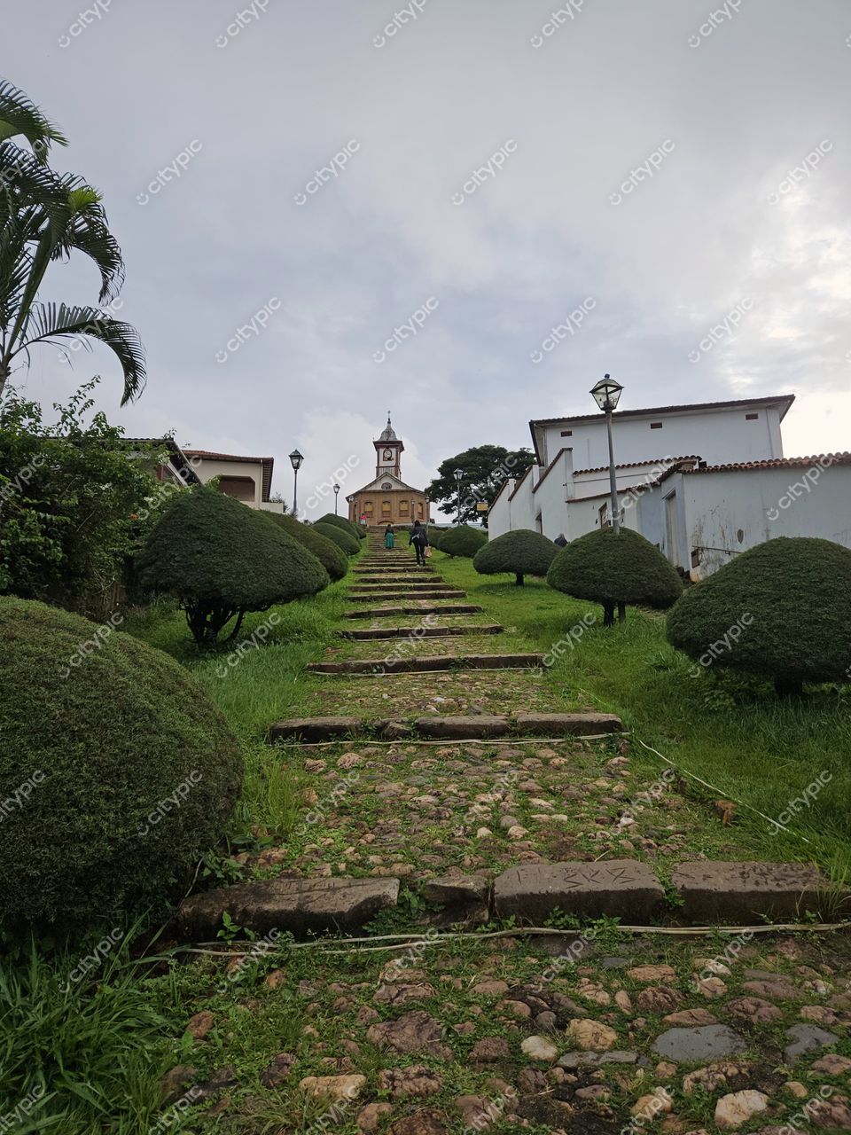 Serro, Minas Gerais caminho de pedras em área rural com grama verde e construção com torre sob céu nublado