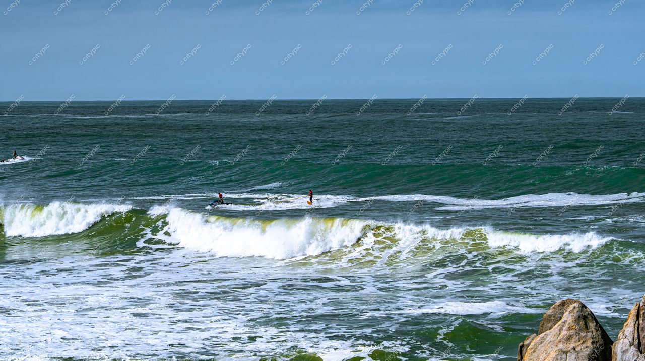 Lagoinha do Leste em Florianópolis com surfistas nas ondas grandes e rochedos à direita sob céu nublado