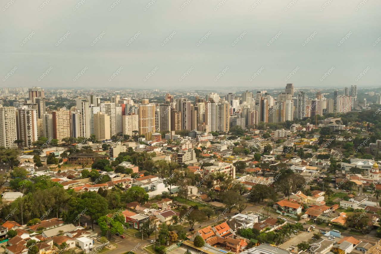 Curitiba, Paraná vista panorâmica com casas de um andar, vegetação densa e arranha-céus sob céu nublado