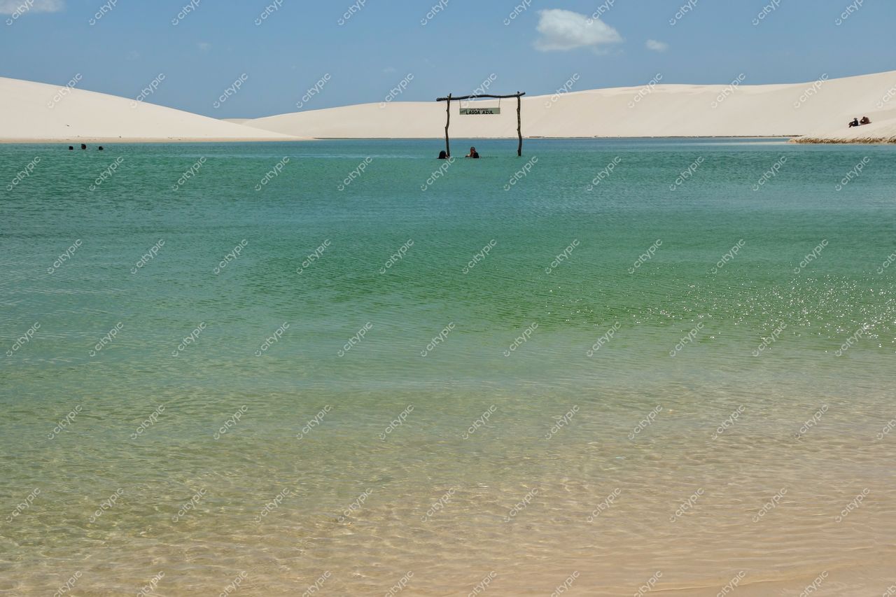 Lençóis Maranhenses, Maranhão, lagoa de águas transparentes com pessoas e dunas brancas sob céu claro e ensolarado