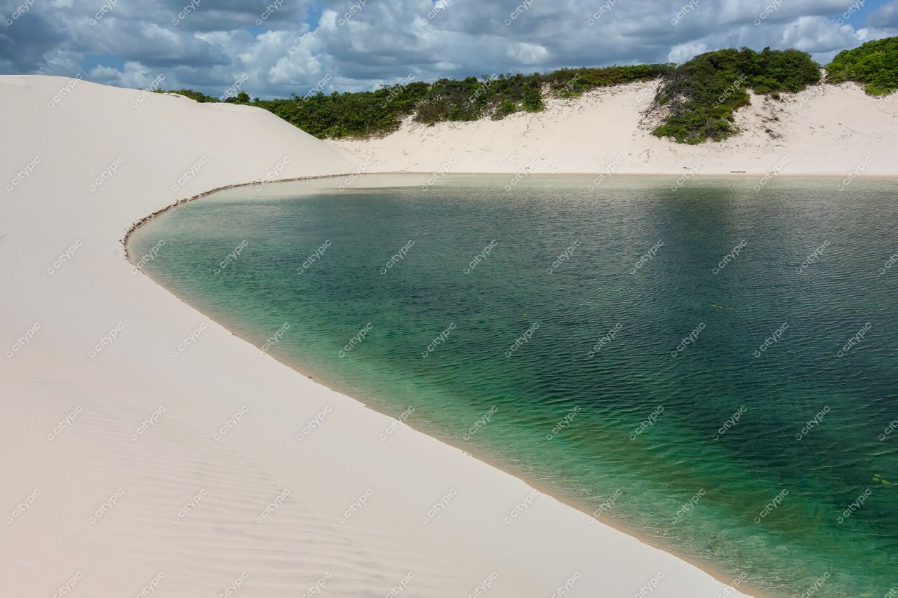 Lençóis Maranhenses, Maranhão, lago cristalino cercado por dunas brancas e vegetação rasteira sob céu parcialmente nublado