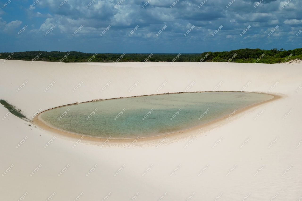 Lençóis Maranhenses, Maranhão, lagoa esverdeada entre dunas brancas sob céu nublado com vegetação ao fundo