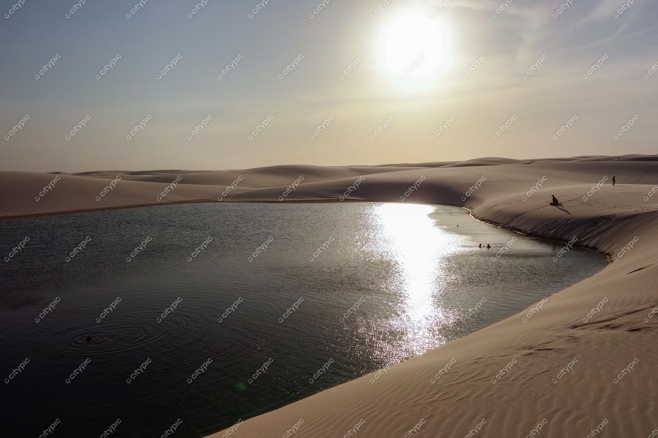 Lençóis Maranhenses, Maranhão, lagoa tranquila refletindo o sol ao entardecer com figuras humanas no cenário sereno