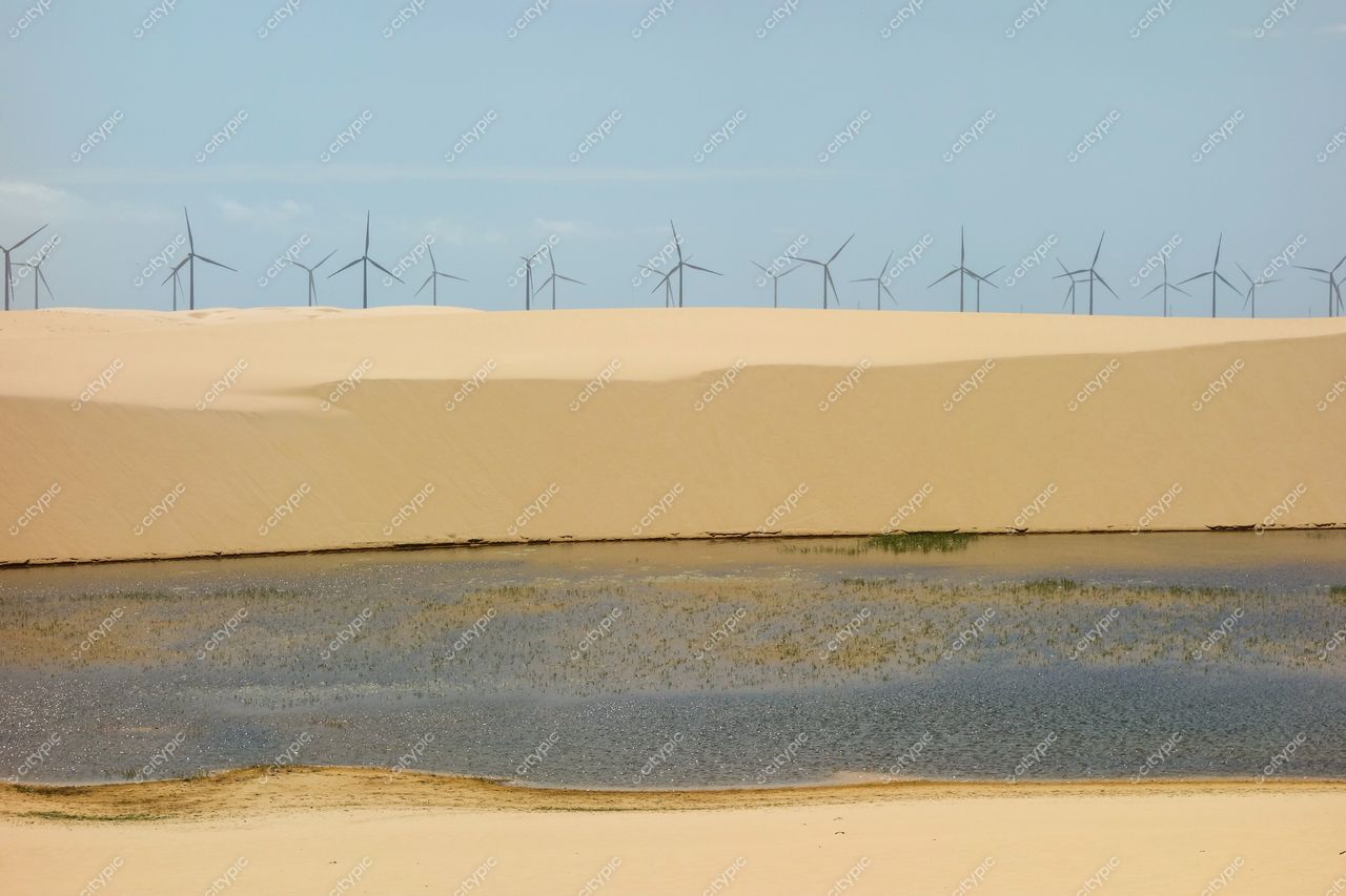 Atins, Maranhão com dunas amarelas e lago em primeiro plano, turbinas eólicas ao fundo sob céu claro