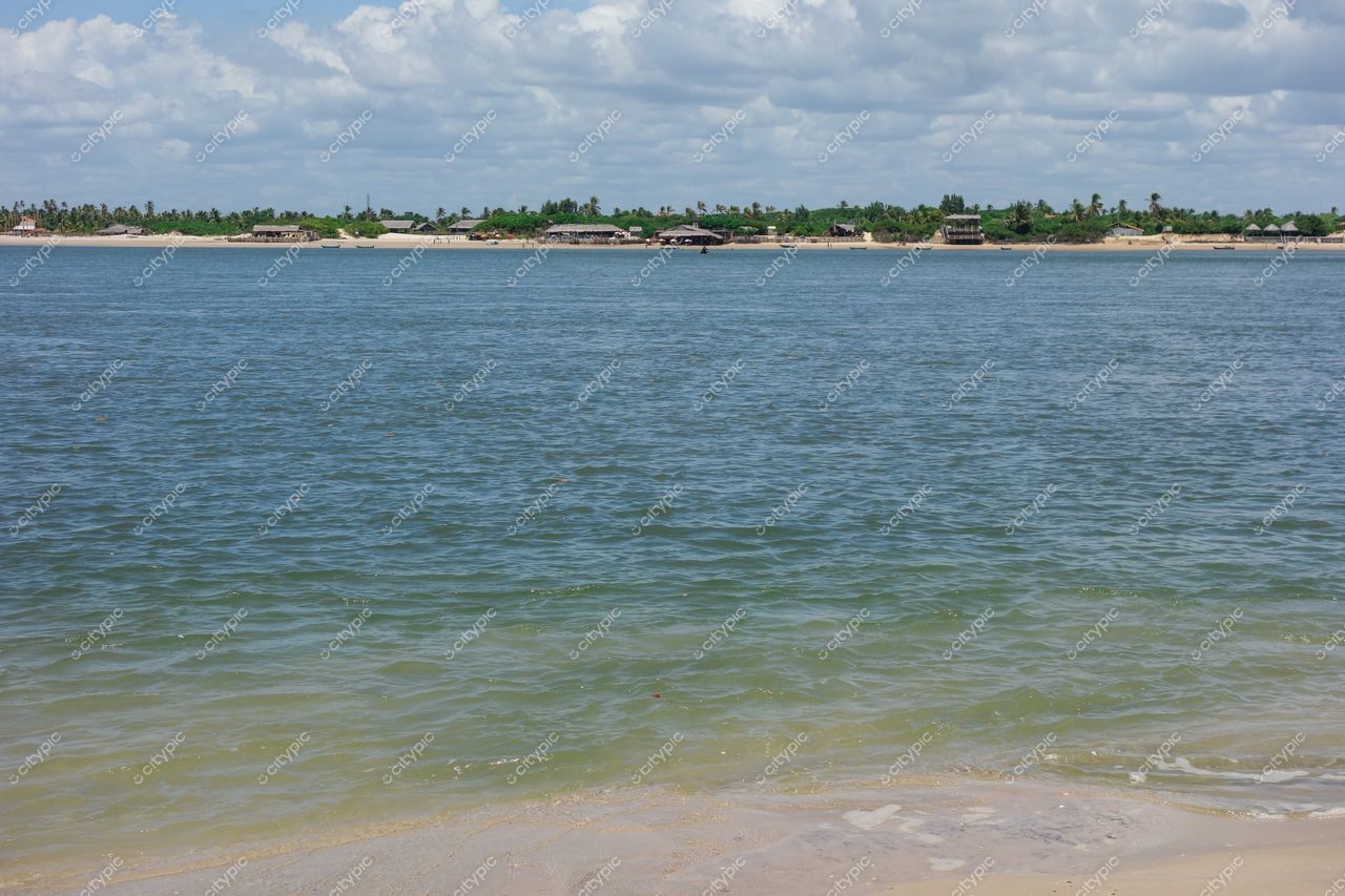 Atins, Maranhão, paisagem serena com rio calmo, casebres de palha e vegetação exuberante sob céu nublado