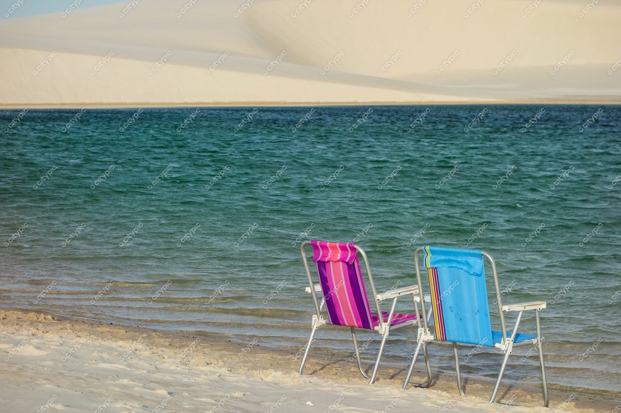 Lençóis Maranhenses, Maranhão, cadeiras de praia rosa e azul à beira de lagoa com dunas brancas ao fundo