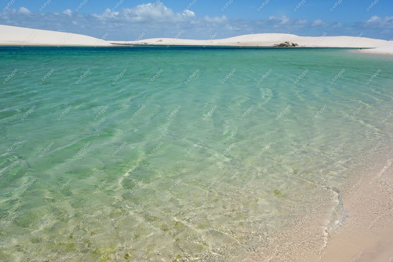 Lençóis Maranhenses, Maranhão, lagoa cristalina em dia claro com céu parcialmente nublado e areia branca ao fundo