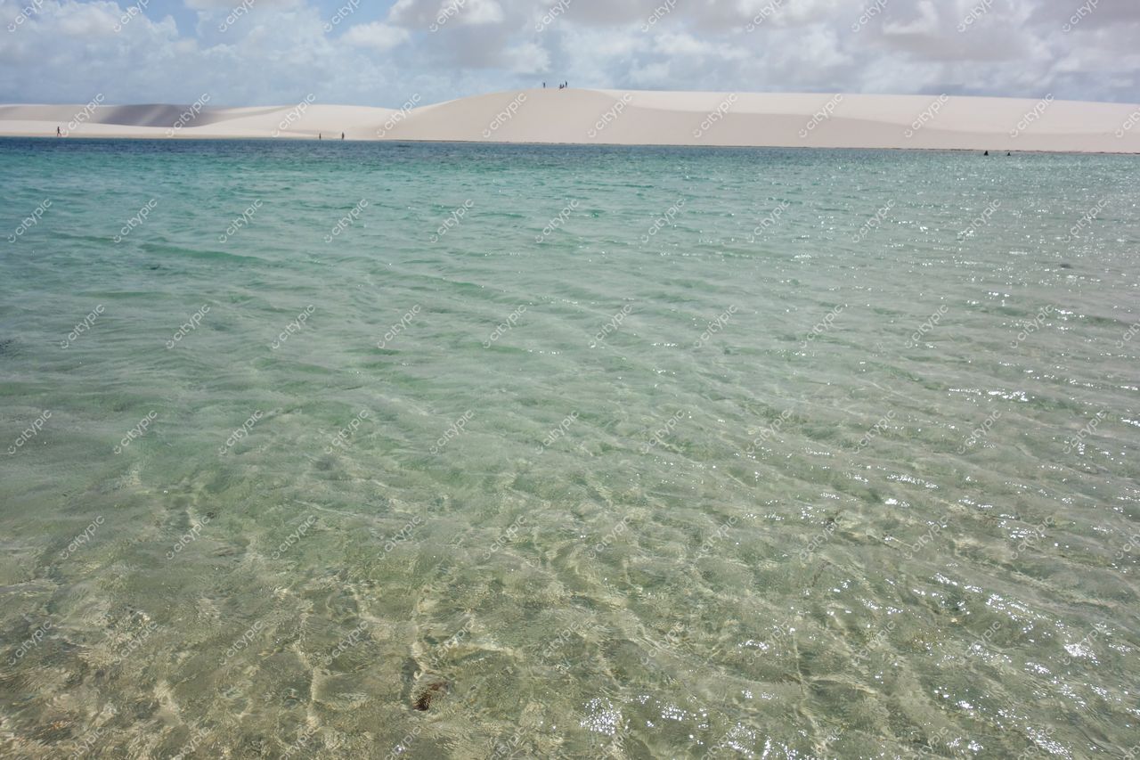 Lençóis Maranhenses, Maranhão, lagoa cristalina com tonalidades azul e verde, dunas brancas e céu parcialmente nublado