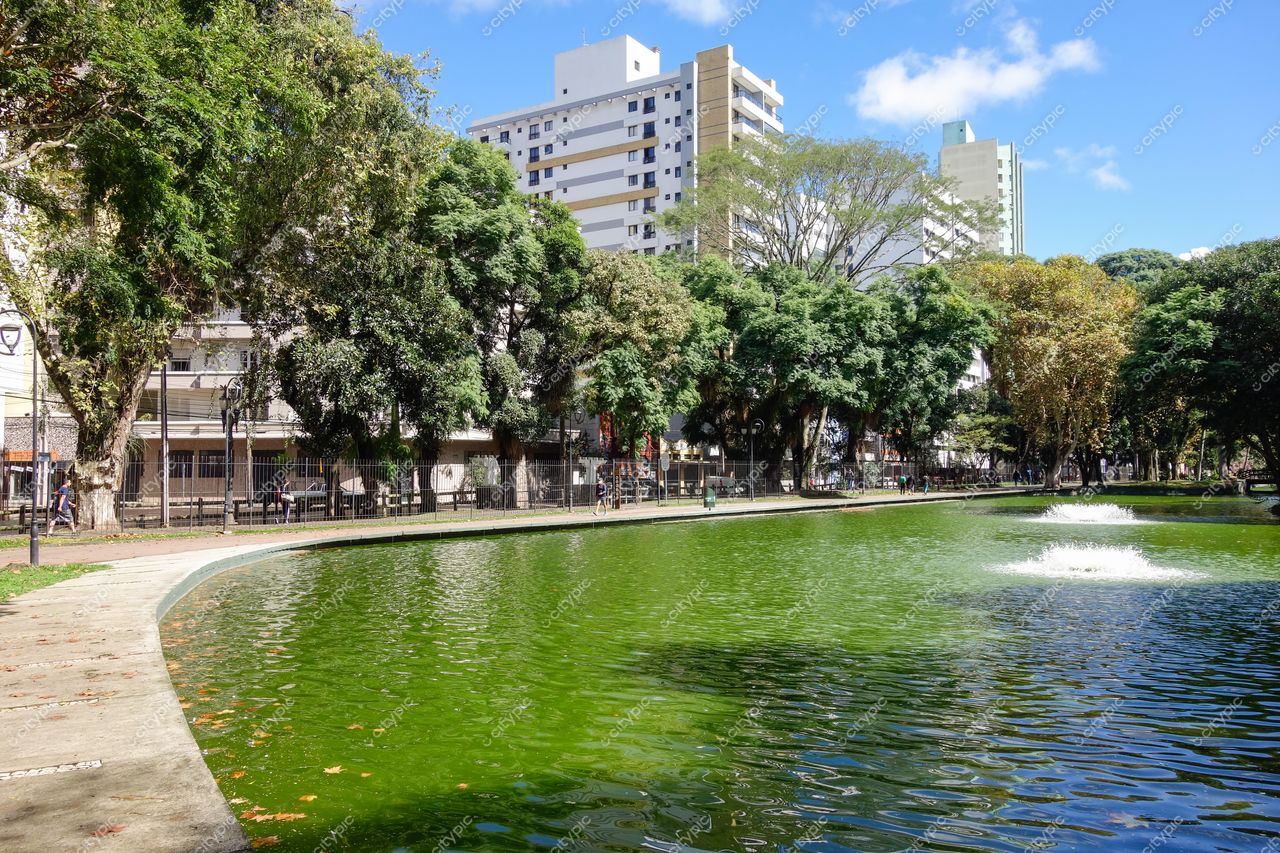 Curitiba, Paraná parque urbano com lago verde e chafarizes em dia ensolarado cercado por edifícios e vegetação variada