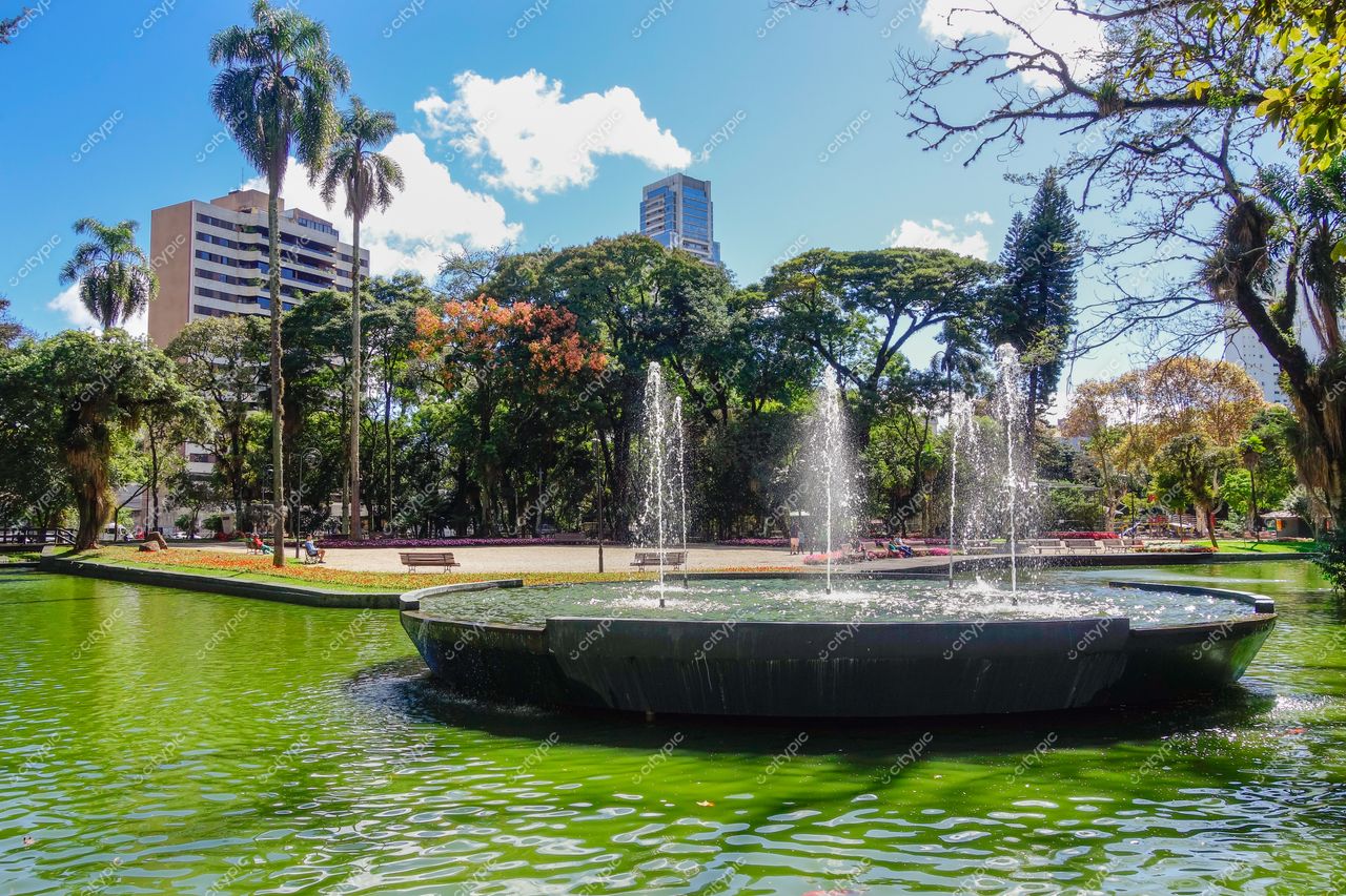 Curitiba, Paraná, parque com fonte circular em lago verde, prédios altos ao fundo e vegetação densa iluminada pelo sol