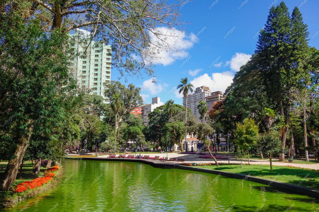 Curitiba, Paraná lago verde refletindo céu azul com flores coloridas e edifícios altos ao fundo em dia ensolarado