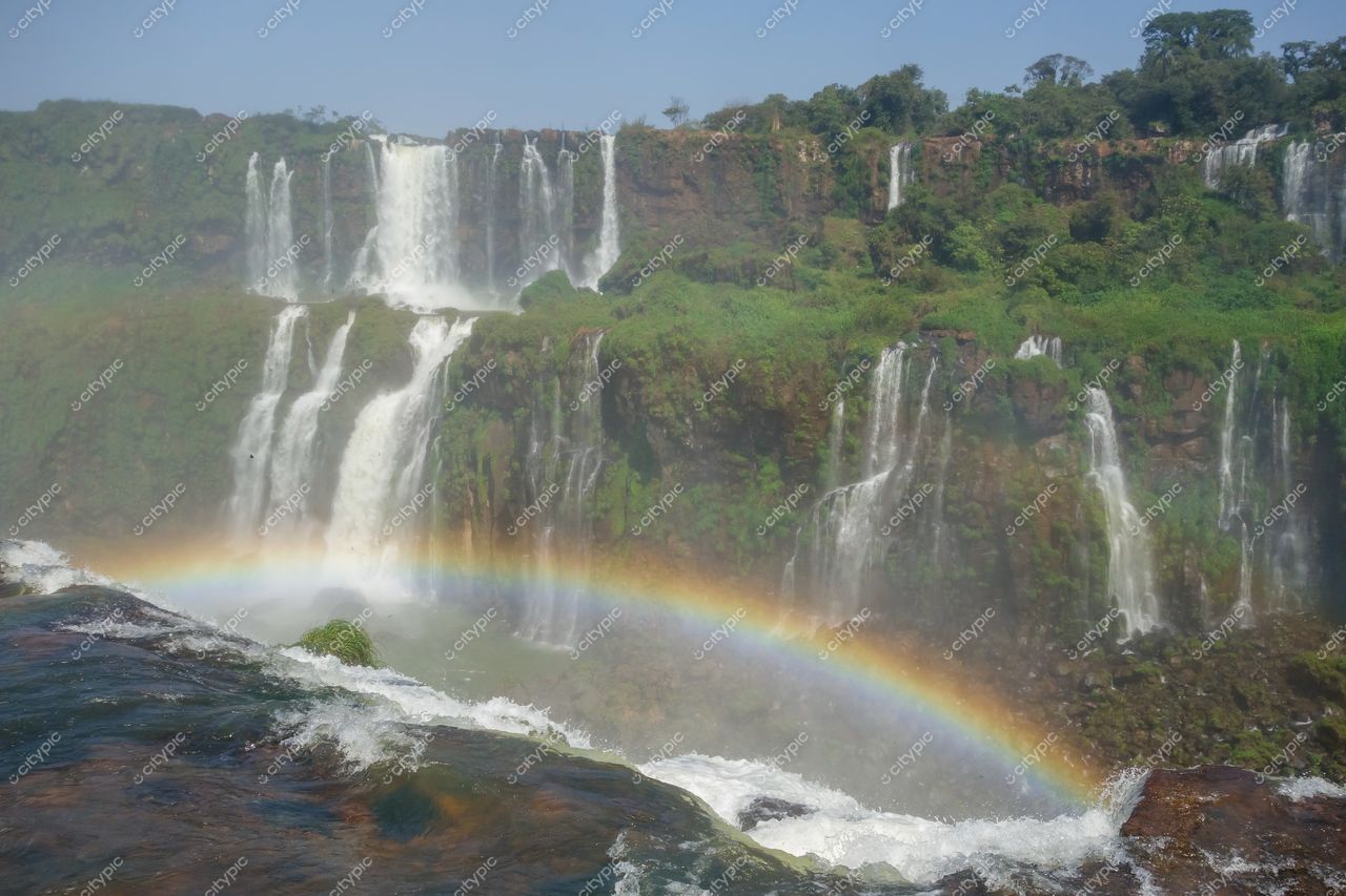 Foz do Iguaçu, Paraná, Cataratas com névoa e arco-íris em dia ensolarado entre vegetação exuberante e relevo verdejante