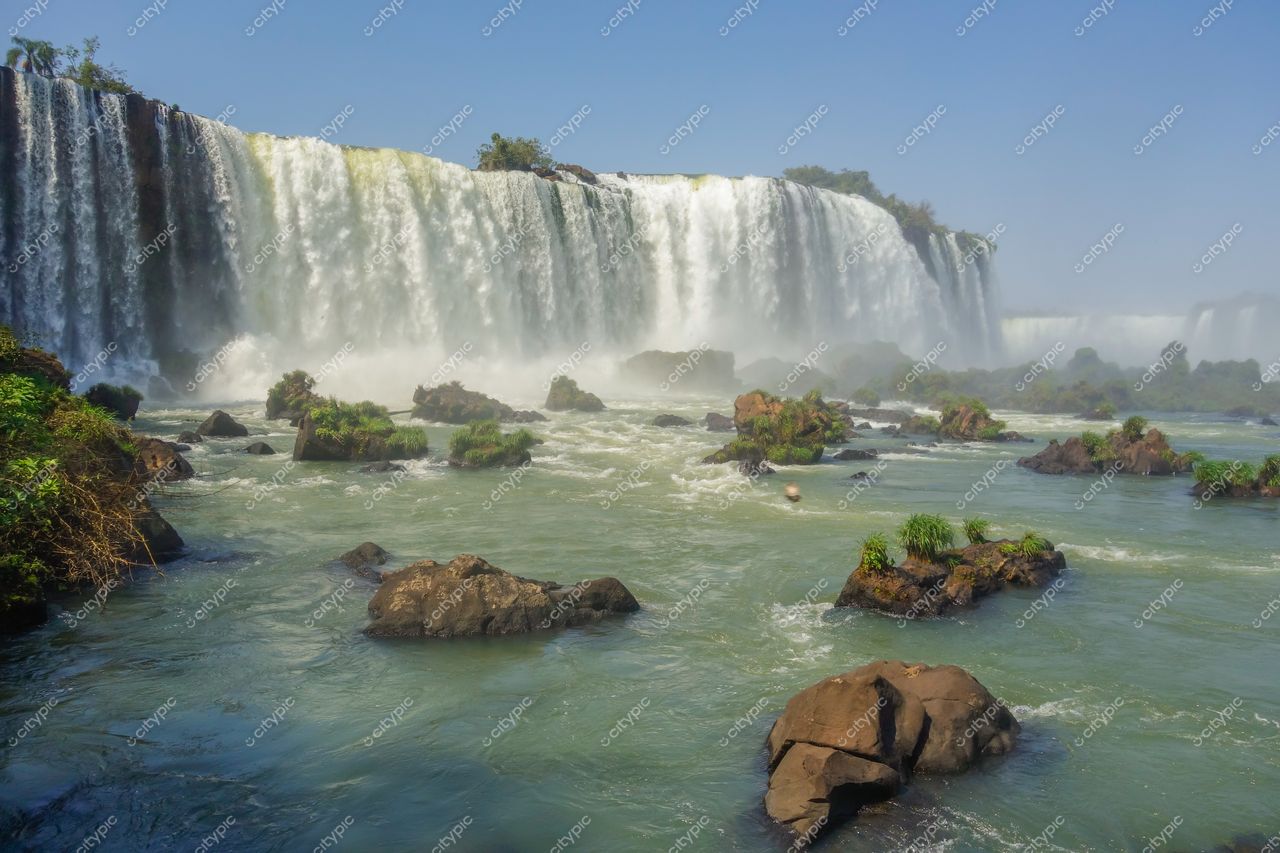 Foz do Iguaçu, Paraná, Cataratas com intenso fluxo de água e vegetação rasteira sob céu limpo e iluminado
