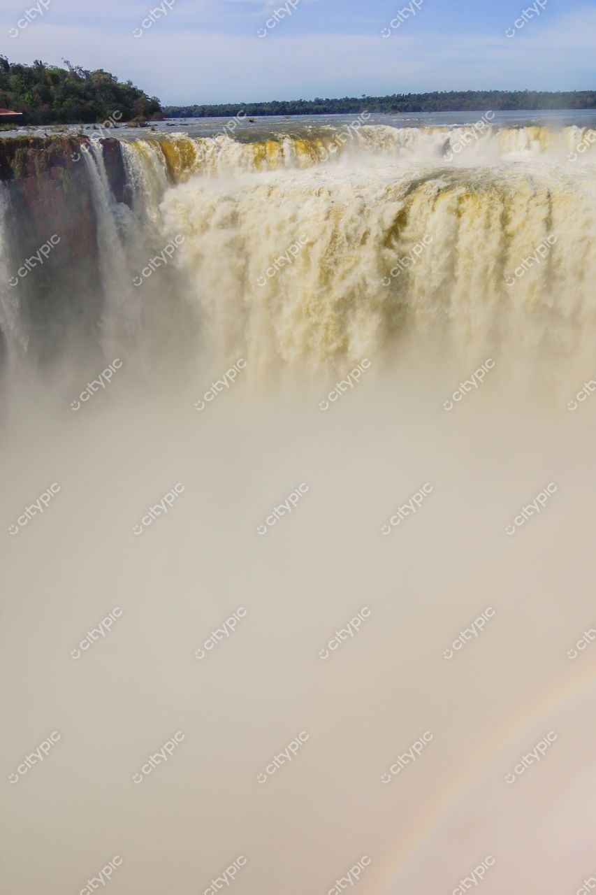Foz do Iguaçu, Paraná, vista das Cataratas com intenso volume de água e arco-íris sob céu azul e nuvens