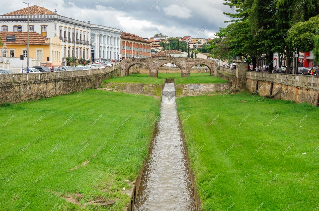 São João del Rei, Minas Gerais, canal de água entre gramados verdes e construções históricas sob céu nublado