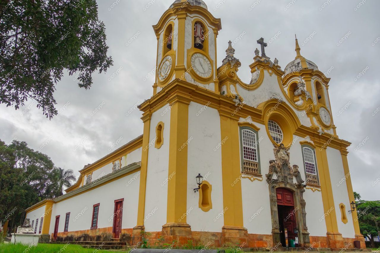 Tiradentes, Minas Gerais, igreja colonial com detalhes em amarelo sob céu nublado e vegetação ao redor