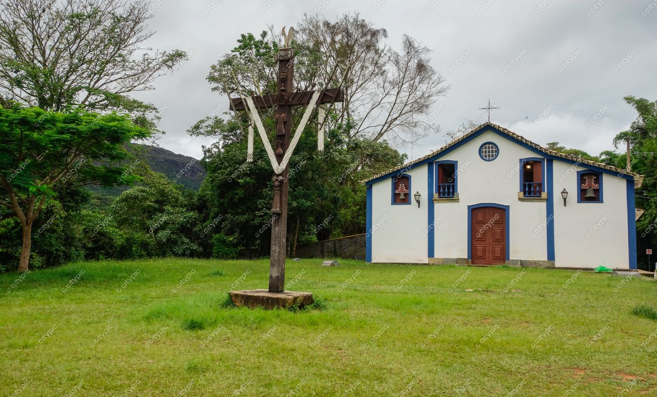 Tiradentes, Minas Gerais, igreja colonial com cruz de madeira em gramado verde sob céu nublado e vegetação densa