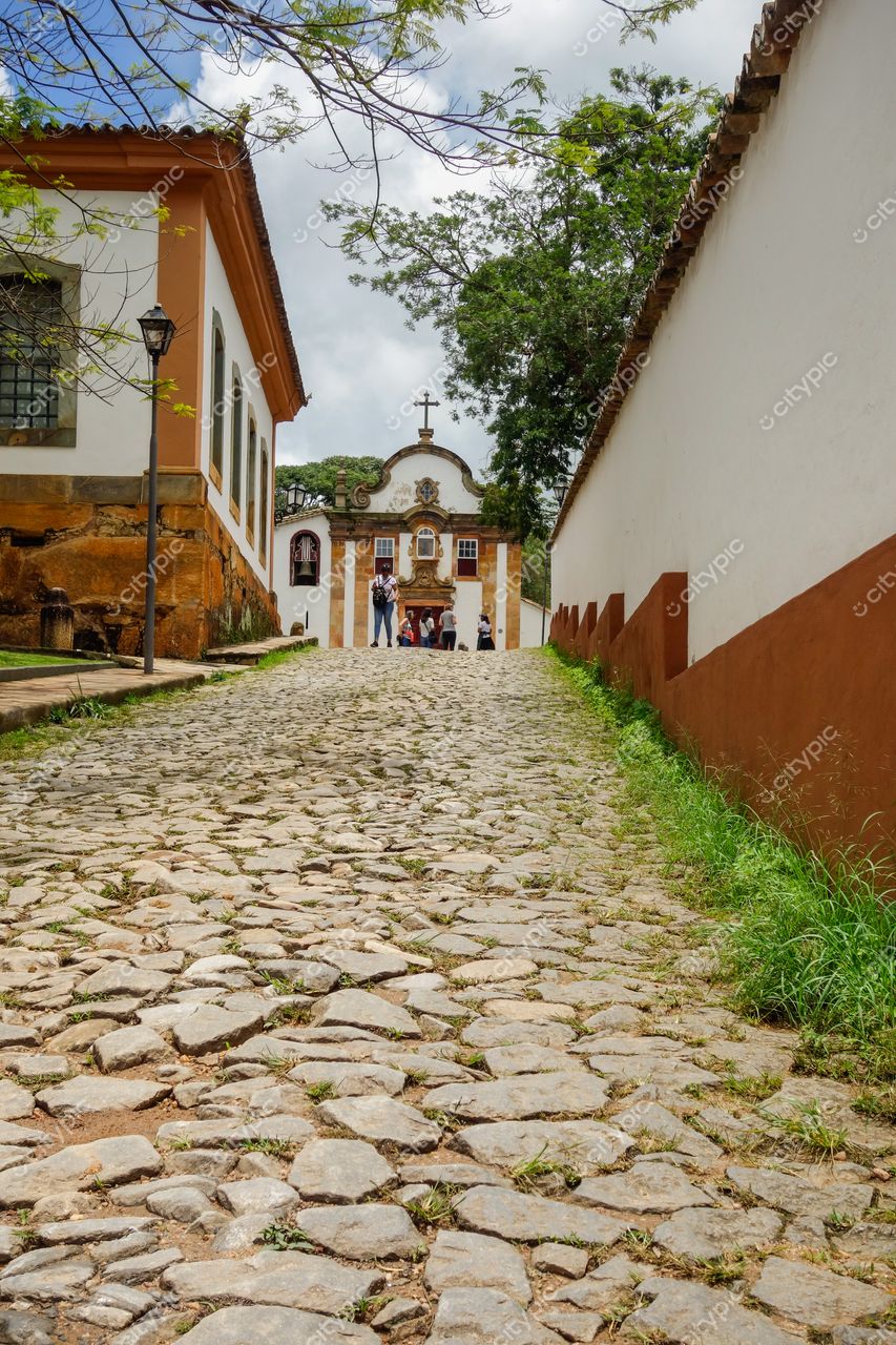 Tiradentes, Minas Gerais, rua de paralelepípedos com casas coloniais claras e igreja ao fundo em dia ensolarado