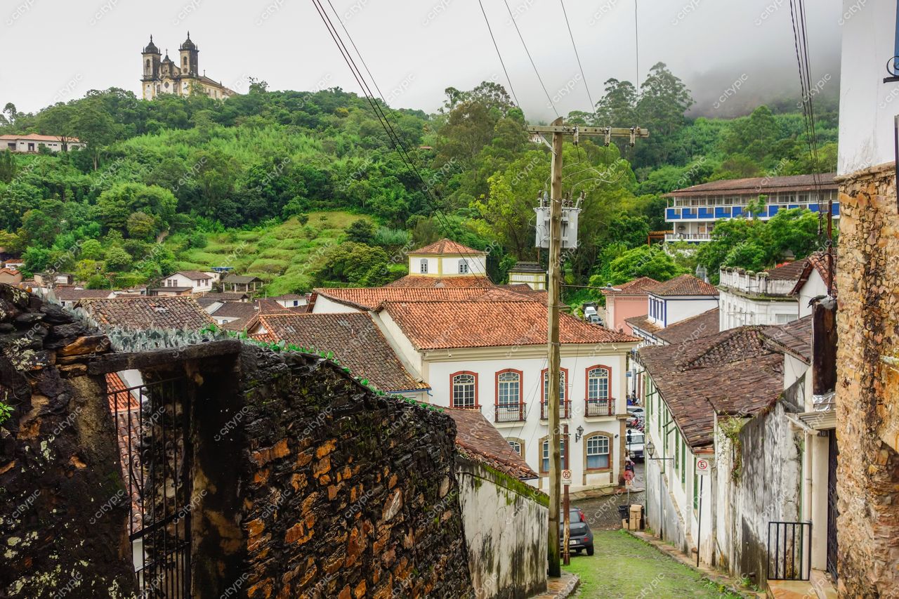 Ouro Preto, Minas Gerais com vista panorâmica de casas coloridas e igrejinha em morro sob nuvens e vegetação densa