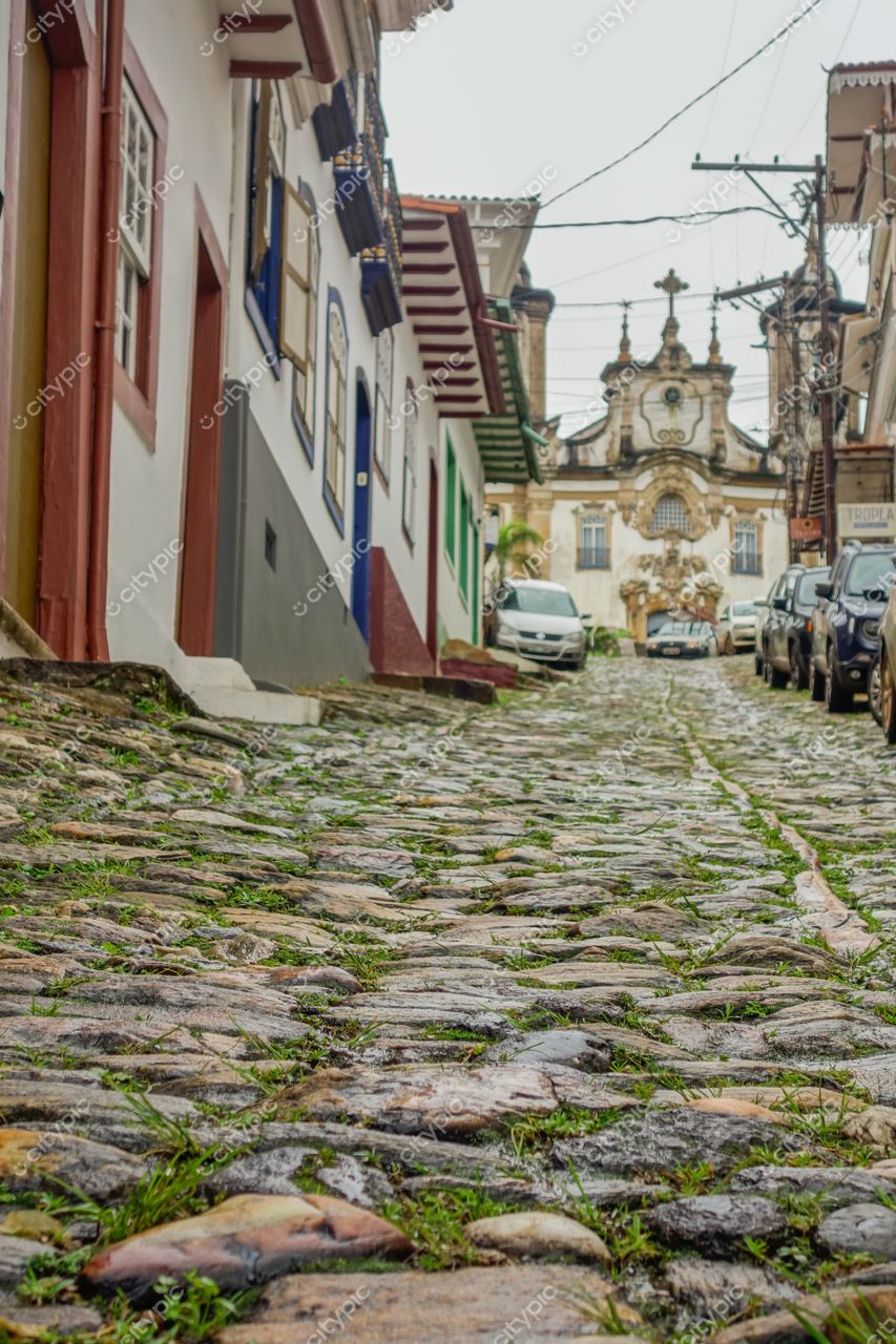 Ouro Preto, Minas Gerais, rua de paralelepípedos com casas coloridas e igreja barroca sob céu nublado e vegetação
