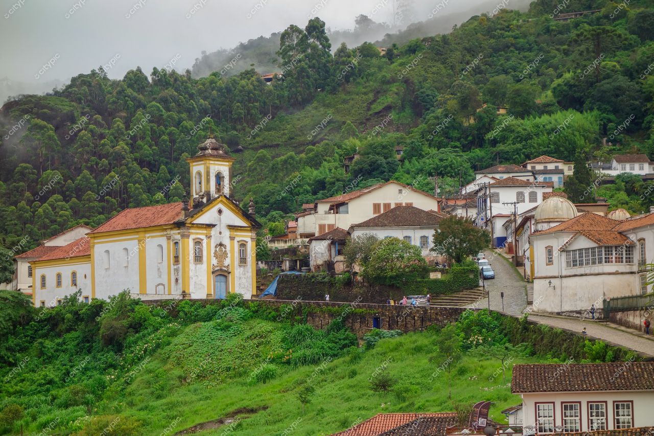 Ouro Preto, Minas Gerais, igreja colonial branca e amarela com vegetação densa em dia nublado e ruas de paralelepípedo