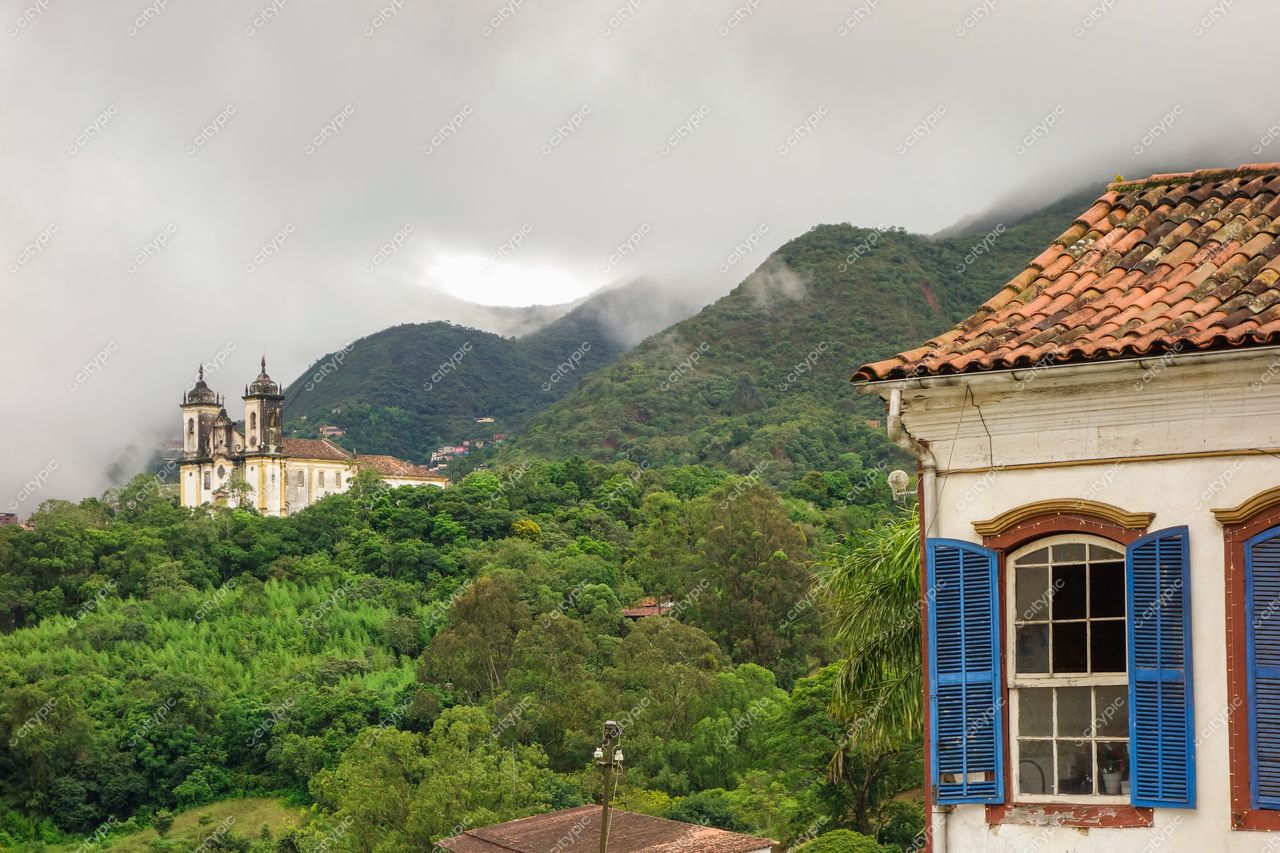 Ouro Preto, Minas Gerais em dia nublado com casa de janelas azuis e montanhas cobertas por neblina e vegetação