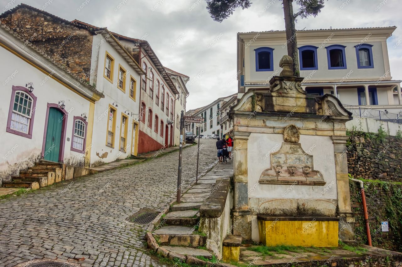 Ouro Preto, Minas Gerais, rua de paralelepípedo com casas coloniais coloridas e fonte ornamental sob céu nublado