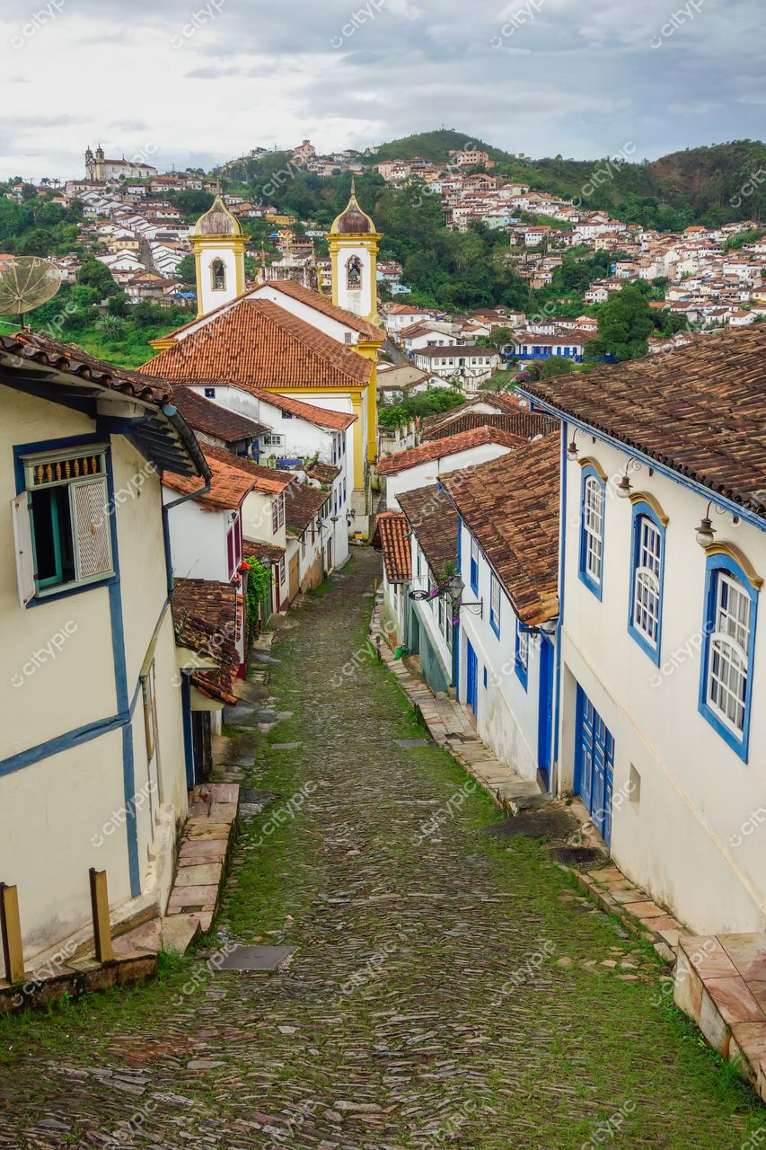 Ouro Preto, Minas Gerais, rua de paralelepípedo com casas coloniais azuis e brancas sob céu nublado e vegetação