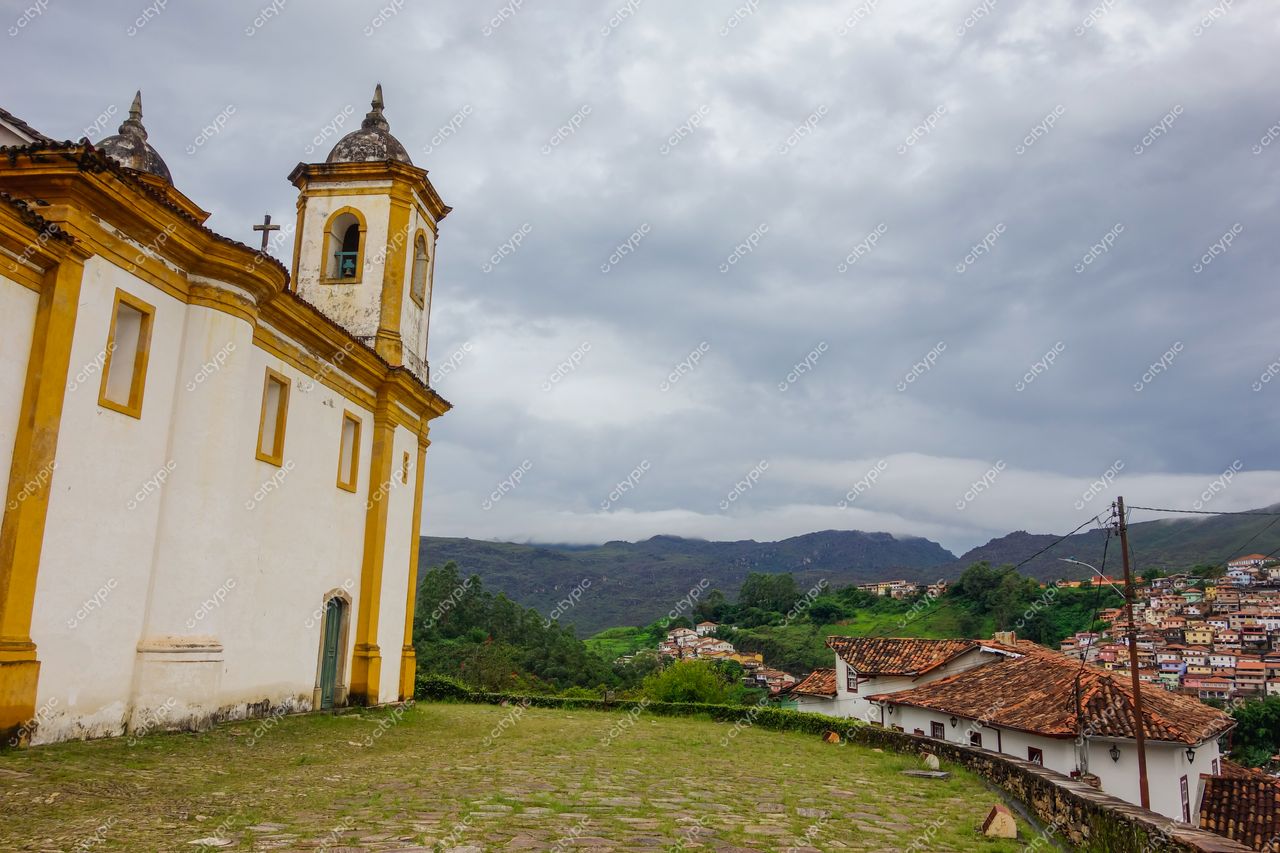 Ouro Preto, Minas Gerais, vista de igreja colonial branca com detalhes amarelos em dia nublado e vegetação montanhosa ao fundo