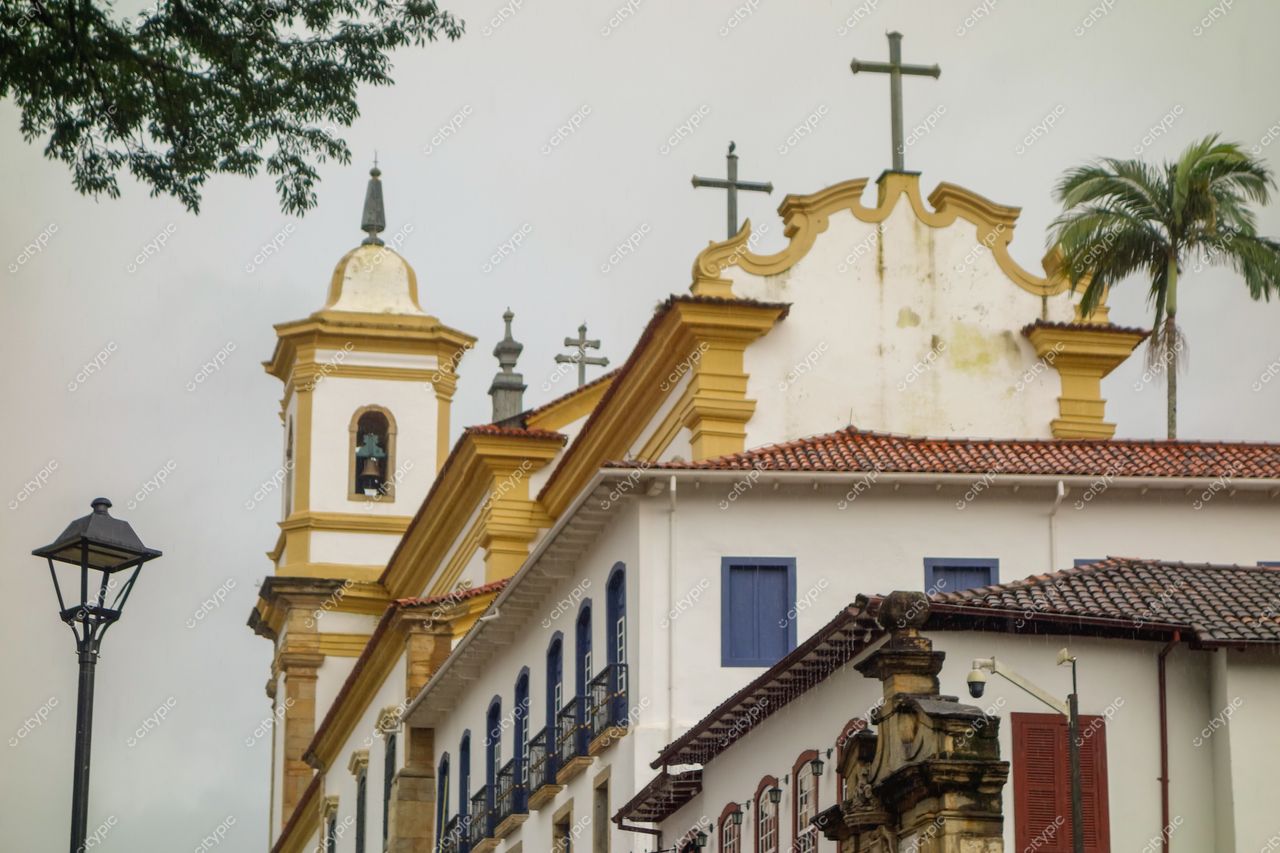 Mariana, Minas Gerais, edifício religioso com paredes brancas e amarelas sob céu nublado e palmeira ao lado
