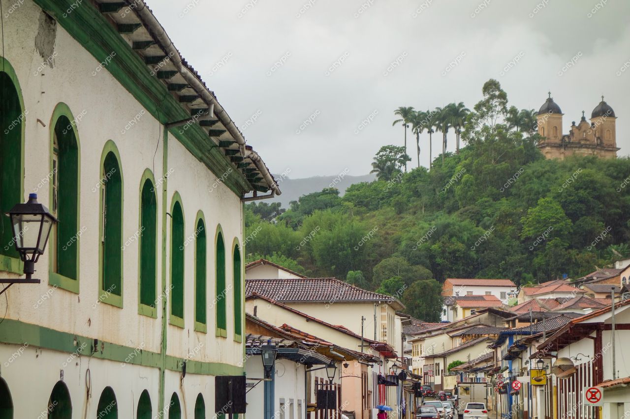 Mariana, Minas Gerais, rua com edifício colonial verde em dia nublado e colina com construção religiosa ao fundo