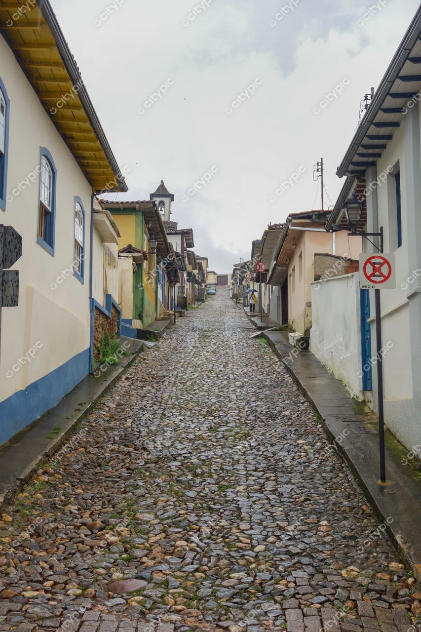 Mariana, Minas Gerais, rua de paralelepípedos com casas coloniais coloridas sob céu nublado e vegetação lateral