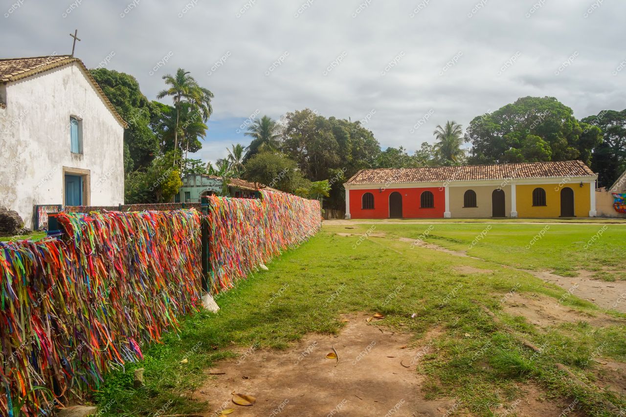 Porto Seguro, Bahia, cerca colorida de fitas em gramado com casa branca e edifício colonial sob céu nublado
